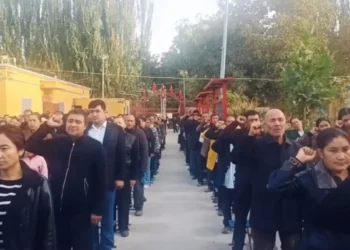 People standing in two parallel lines outdoors, many raising their right hands to their heads in a salute during a ceremony.