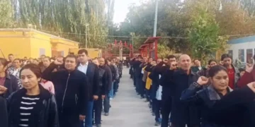 People standing in two parallel lines outdoors, many raising their right hands to their heads in a salute during a ceremony.