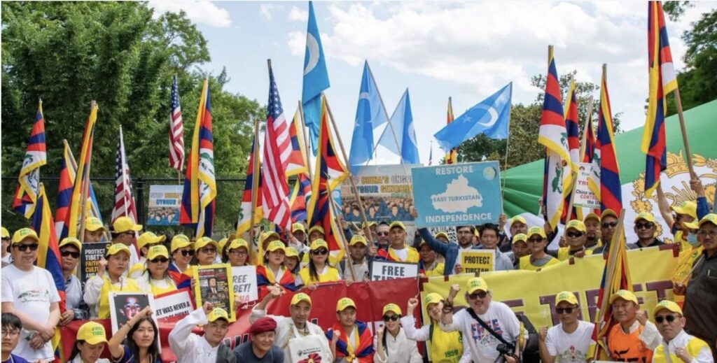 Screenshot | The East Turkistan Post Crowd of people at a pro-independence rally holding colorful flags and yellow hats, with banners and signs visible against a blue sky and trees in the background.